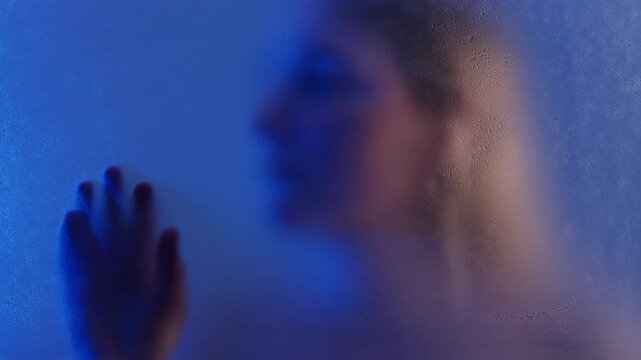 Anonymous blond woman touching wet matte glass in blue light. She is feeling lonely and sad. 