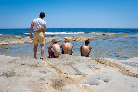 cousins of different ages enjoying a family vacation by the rocky Mediterranean sea during a sunny summer day, with their backs turned, looking out at the sea, wearing funny straw hats