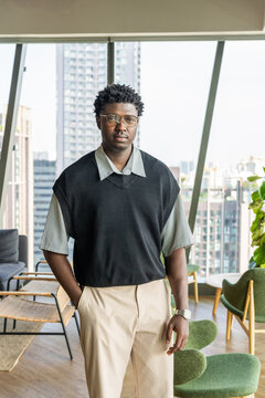 Black Man Stands in Modern Workspace With City View Behind Him