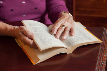 Fototapeta premium An elderly woman's hands rest on the pages of an open book at a wooden table, capturing a quiet moment of reading, memory and leisure in later life.