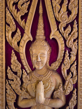 Detailed golden carving of a traditional Thai deity with hands pressed together in a praying gesture against a red backing.