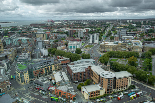 Aerial view captures Liverpool's urban landscape under cloudy skies. Modern and historic buildings mix across the frame. Green spaces and roads connect neighborhoods. River and docks show distant mari