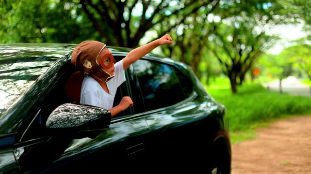 Smiling young Asian girl wearing a retro aviator hat leaning out of a modern car window, raising her arm joyfully towards the beautiful blue sky with freedom under green tree canopy.