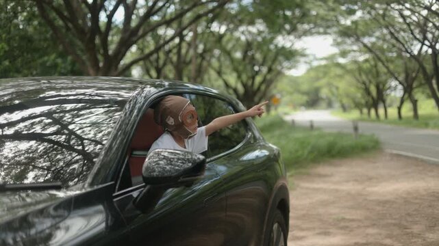 Joyful little Asian girl wearing a vintage aviator hat leaning out of a black car window, extending her hand forward like flying with a bright cheerful smile in green nature.