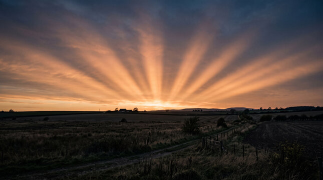 Radiant golden sun rays shining through dramatic clouds over a vast rural landscape during sunset