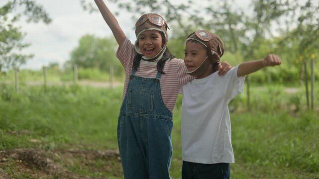 Joyful little Asian children wearing vintage aviator pilot hats standing together outdoors on green field, raising their hands triumphantly with bright smiles during summer day.