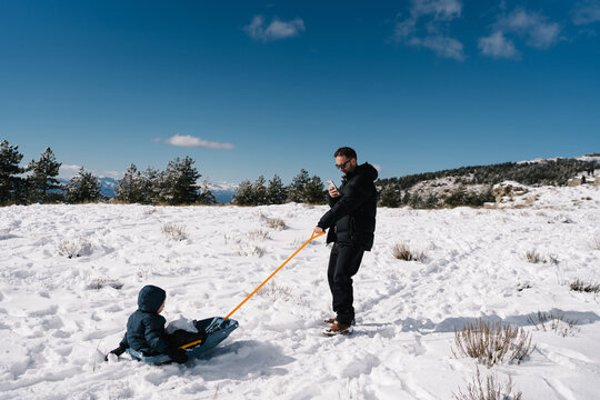 Father pulling child on sled while checking smartphone