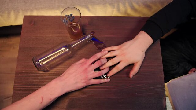 Top table view of small wooden tabletop with empty bottle on side, spilled drink and glass. A male drunk hand reaches for key fobs, but a sober hand takes away the car keys to prevent a drunk driving