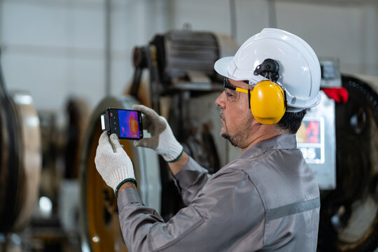 professional industrial engineer in safety gears using smartphone connected thermal imaging camera to detect heat signatures and perform predictive maintenance in factory