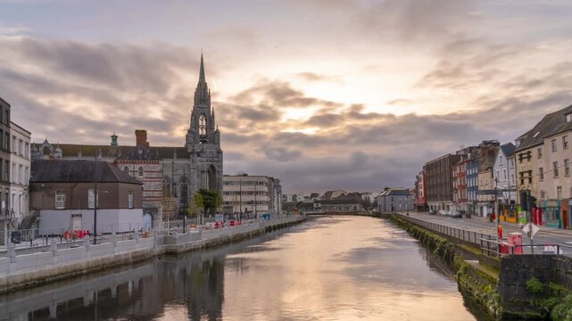 A view of the River Lee in Cork, Ireland, with St. Fin Barre's Cathedral in the background. The river is calm, reflecting the cloudy sky and surrounding buildings.
