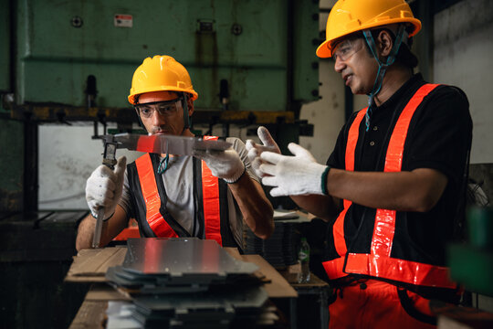 Quality Inspectors Measuring a Manufactured Metal Component with a Caliper Beside Stacked Parts and Industrial Machinery in a Factory Workshop