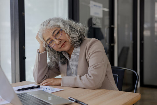 Tired Senior Businesswoman Resting Head on Hand at Office Desk with Laptop and Documents