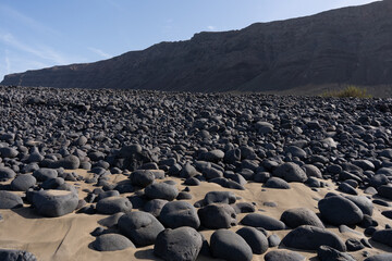 Obraz premium Volcanic black pebbles and golden sand covering beach with arid mountains under blue sky, Canary Islands destination