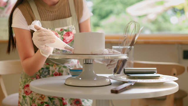 Young Asian woman applying frosting to round cake on revolving stand in bright kitchen with window view