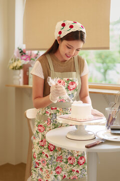 Young Asian woman decorating white cake with piping bag in sunny home kitchen wearing floral apron