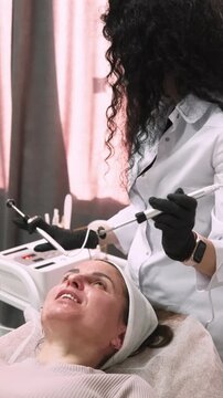 Vertical video. Microcurrent rejuvenation. Female skin. Beauty master in black gloves using professional device for facial lifting procedure.