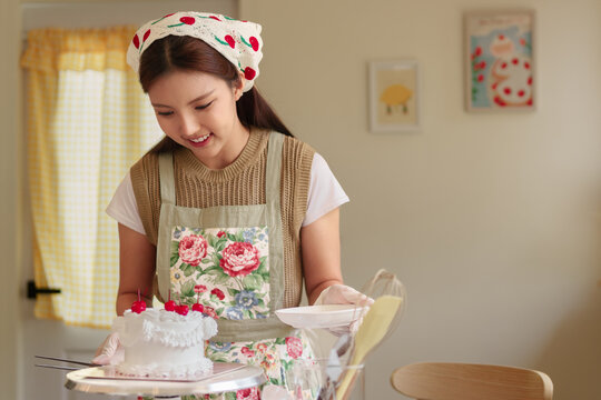 Young Asian woman admiring decorated white cake with cherries in vintage kitchen with copy space