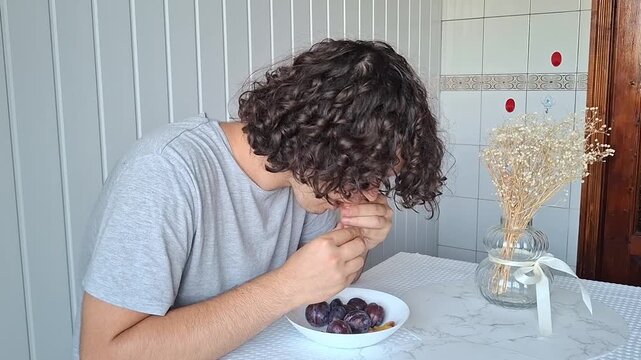 Young man eats a bowl of fruit at a kitchen table in a simple home