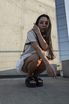 Low-angle shot of a stylish young Asian woman crouching on an urban rooftop, posing for the camera in casual attire and platform sandals against a concrete and glass background.