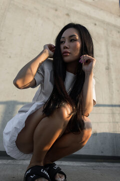 Low-angle portrait of a pensive young Asian woman sits on an urban rooftop, looking away while adjusting her long hair during sunset against a textured concrete wall backdrop. Pretty sexy lady