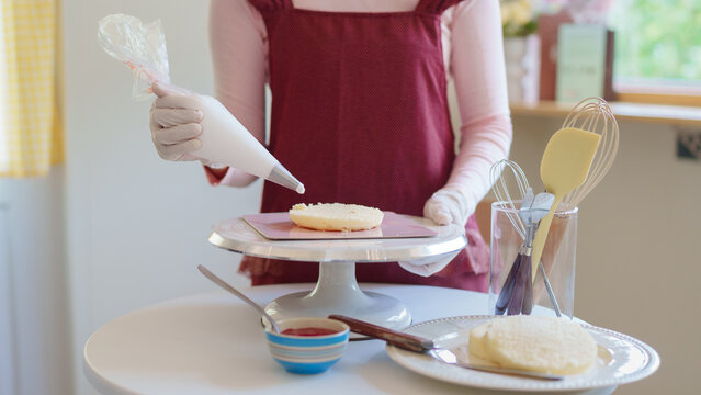 Woman in burgundy apron piping frosting onto sugar cookies using pastry bag in bright home kitchen