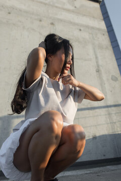 young Asian woman in casual clothes squats by a concrete wall, adjusting her long hair under warm afternoon sunlight. Perfect for urban fashion, youth culture, and lifestyle stock imagery.