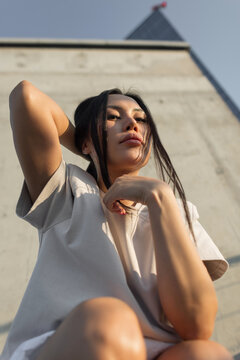 Low angle portrait of a confident young Asian woman looking at the camera against a concrete wall in warm afternoon sunlight, holding her long dark hair. Great for urban fashion.