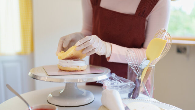 Asian woman baker assembling cream layer cake on stand in bright home kitchen