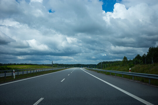 First-person perspective from a car driving on an empty asphalt highway. A modern expressway with a bridge overpass under a dramatic, cloudy stormy sky. No other traffic.