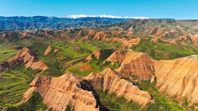 Aerial view of colorful red clay hills and green valleys against a background of snow capped mountains under blue sky in Xinjiang, China.