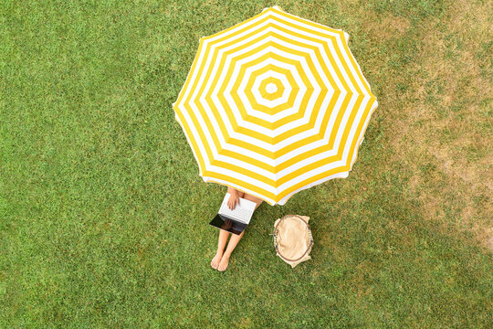 Woman in a white bikini with basket for picnic sitting on deck chair under yellow umbrella  on the green grass sunbathes at summer day. Top view, drone, aerial view.
