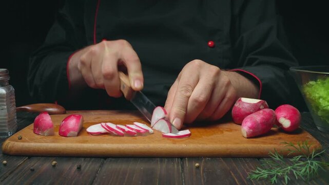 Chef in black uniform slices fresh radishes on a wooden cutting board, with vibrant vegetables and kitchen tools visible in the background
