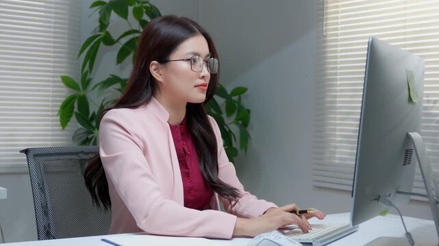 Professional asian businesswoman with glasses typing on a computer keyboard and smiling in a modern office environment