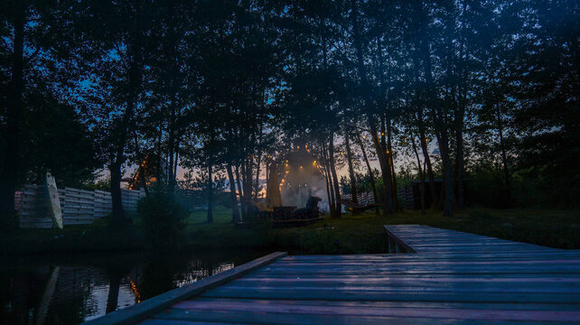 Twilight scene with a wooden pier, campfire, and string lights in a forest clearing