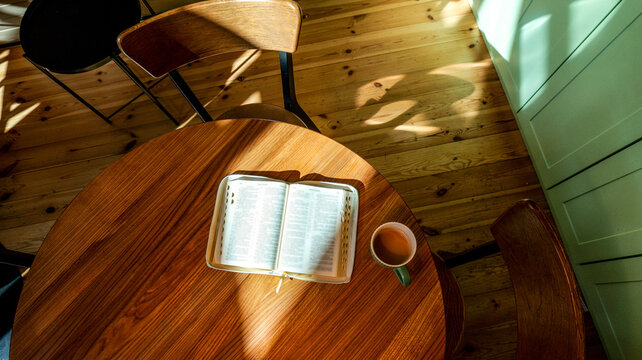 Open bible and coffee cup on wooden table with sunlight streaming through window