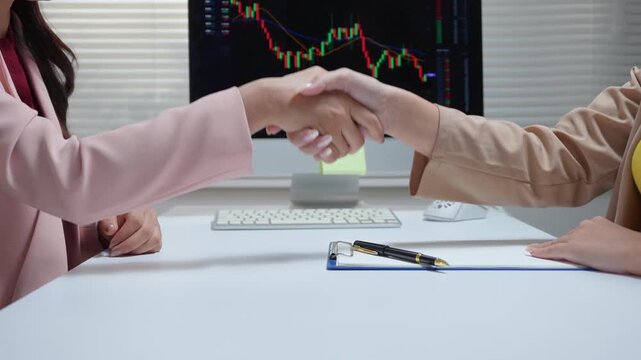 Businesswomen shaking hands in agreement over stock market data on a computer screen in the office
