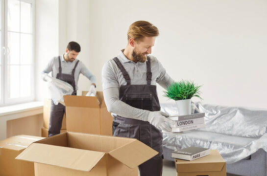 Team of two young confident cheerful professional workers of moving service in overalls packing and delivering cardboard boxes in new apartment. Move, moving day and relocation concept