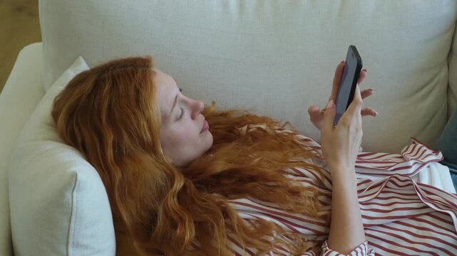 A young woman with long, red hair lies comfortably on a white couch, engrossed in scrolling through content on her smartphone. She appears relaxed and enjoying her digital interaction.