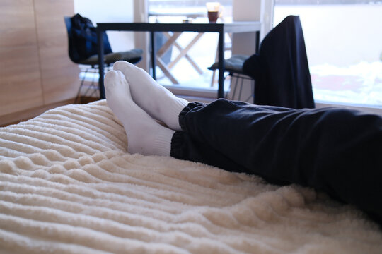 Cozy morning indoors as feet clad in plush white socks relax upon a warm fuzzy blanket while snow glistens outside the window pane during a serene winter day in comfort.