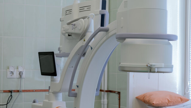 Interventional radiology C-arm fluoroscopy system beside adjustable patient table with orange pillow in clean tiled procedure room under soft clinical lighting