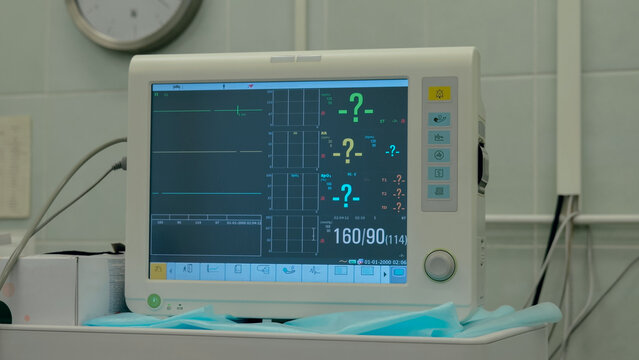 Bedside patient monitor showing flat cardiac waveform and numeric vital readings on wheeled trolley in quiet clinical room with blue surgical drape, tiled wall, soft fluorescent light, and medical