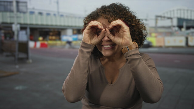 Woman making binocular gesture with hands by eyes on paved street in front of building facade; curiosity exploration.