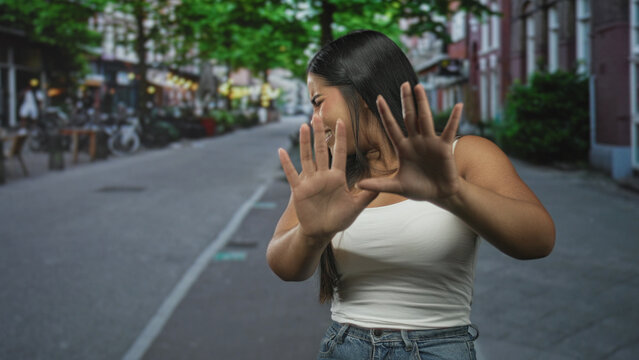 Young hispanic woman with palms facing camera in a stop gesture on a street with storefronts and bicycles; alarm avoidance.