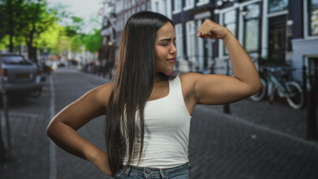 Hispanic woman flexing biceps on a street, fists raised in a confident pose, wearing white tank and jeans against urban buildings and bicycles; confidence empowerment.