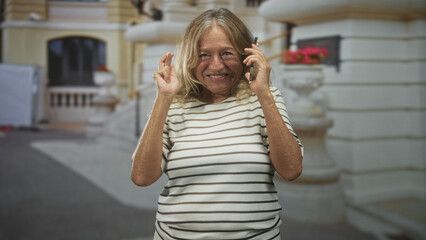 Fototapeta premium Senior blonde woman holding smartphone to ear and crossing fingers while smiling in front of ornate building; joyful celebration.