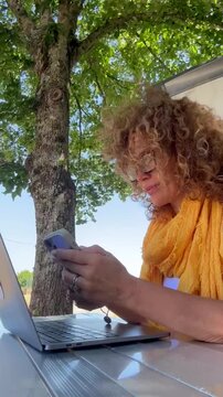 Mature woman with curly hair and glasses works remotely outdoors, focused on her phone and laptop under a tree. This scene embodies modern freelance business, digital communication, and a relaxed, fle