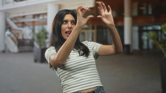Woman framing an imaginary subject with hands and sticking tongue out while miming a camera in an airport concourse; playful focus.