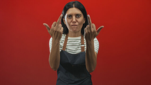 Woman wearing striped shirt and denim apron shows middle fingers with both hands and sticks out tongue in red studio; defiance.