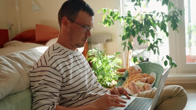 Young single father on paternity leave typing on his laptop while taking care of his newborn baby resting in a bouncer, showcasing a modern work life balance and remote working lifestyle