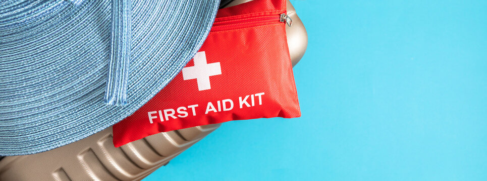 Red first aid kit pouch with straw hat on suitcase over blue background. Medical travel insurance concept, summer vacation health care banner with copy space, safe trip preparation backdrop.